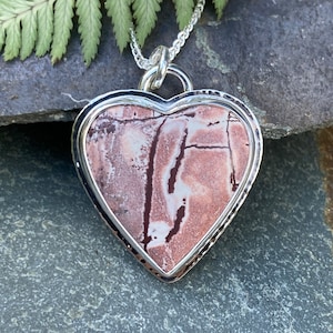 May include: A heart-shaped pendant with a silver frame and chain. The pendant has a stone centre with light brown, white, and dark brown patterns. The pendant is displayed on a grey stone surface.