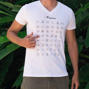 以下が含まれることがあります： 前面に黒いハートと「Companion」という文字がプリントされた白いTシャツ。シャツには、飛行機、家、電話、車、山、自転車、鍵、雲、音符、コーヒーカップなど、小さな黒いアイコンが散りばめられています。