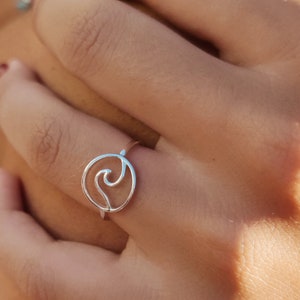 Könnte beinhalten: Ein silberner Ring mit einem Wellenmuster. Der Ring ist minimalistisch gestaltet und hat ein kreisförmiges Band mit einem wellenförmigen Design in der Mitte. Der Ring wird an einem Finger getragen, und das Bild ist bei natürlichem Licht aufgenommen.