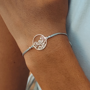 Könnte beinhalten: Ein silberfarbenes Armband mit einem runden Anhänger, der ein ausgeschnittenes Wellenmuster zeigt. Der Anhänger ist an einem dünnen, hellblauen Kordelband befestigt, das um das Handgelenk verläuft und mit kleinen silbernen Perlen fixiert ist.