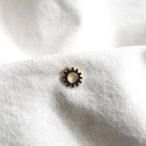 Op de afbeelding: Een gouden zonhanger met een zwart midden, op een witte stoffen achtergrond.