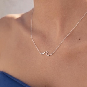 May include: A delicate silver-toned necklace featuring a minimalist wave-shaped pendant centered on a thin, fine-link chain. The metallic wave design is smooth and continuous, resting against the collarbone area with a simple, clean aesthetic.