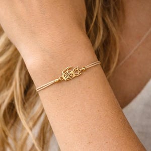 Könnte beinhalten: Ein goldfarbenes Armband mit geometrischem Design, auf einem beigefarbenen Kordelband. Das Armband wird an einem Arm getragen und zeigt die filigranen Details des Schmucks. Das Design scheint eine stilisierte Darstellung eines Vogels oder einer abstrakten Form zu sein.