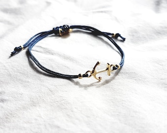 May include: A navy blue cord bracelet featuring a polished gold-tone metal anchor charm at the center. The bracelet includes small gold-tone metal beads along the cord and an adjustable sliding knot closure with knotted ends for a secure fit.