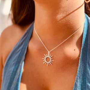 May include: A silver sunburst pendant necklace. The pendant features a circular center with radiating spikes. The necklace is worn on a delicate chain against a blue top. The sun pendant is a popular jewelry item.