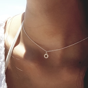 Pode incluir: Um delicado colar de prata com um pequeno pingente em forma de sol. O pingente tem um design detalhado e pontiagudo. O colar é usado perto do pescoço, criando um visual minimalista e elegante. A corrente é fina e sutil.