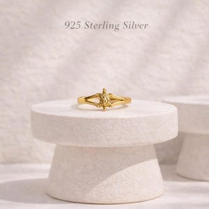 Könnte beinhalten: Ein goldfarbener Ring mit einem kleinen, schildkrötenförmigen Mittelstück. Das Band besteht aus 925er Sterlingsilber. Der Ring ist mittig auf einem runden Steinsockel platziert. Der Text 925 Sterling Silver ist über dem Ring sichtbar.