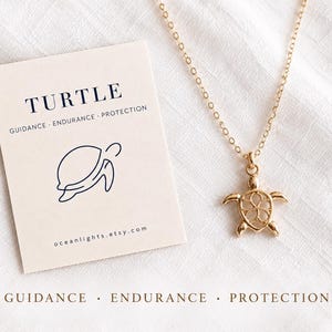 Op de afbeelding: Een goudkleurige ketting met een kleine schildpadhanger aan een fijne ketting. Daarnaast ligt een rechthoekige kaart met de tekst TURTLE, GUIDANCE, ENDURANCE, PROTECTION, een lijntekening van een schildpad en de URL oceanlights.etsy.com.