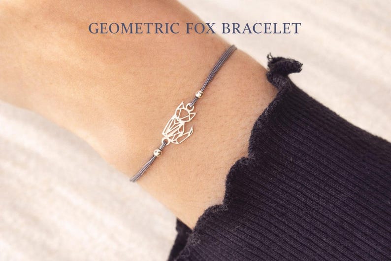 May include: A delicate bracelet featuring a silver-toned geometric fox charm on a thin grey cord. The design includes two small metallic beads flanking the fox. The text GEOMETRIC FOX BRACELET is printed in the upper portion of the image.