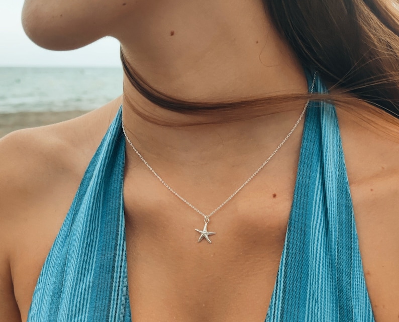 May include: A silver starfish pendant necklace. The necklace features a delicate chain and a small, detailed starfish charm. The background shows a blue and white striped halter top and a blurred beach scene.