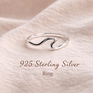 May include: A 925 sterling silver ring featuring a single wave-shaped design element. The band is thin and polished, with the text 925 Sterling Silver Ring printed clearly below the jewellery piece on the fabric surface.