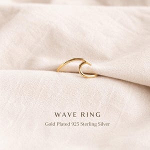 Könnte beinhalten: Ein goldbeschichteter Wellenring aus 925er Sterlingsilber. Der Ring hat die Form einer stilisierten Meereswelle. Der Ring wird auf einem cremefarbenen Stoff präsentiert. Der Text "WAVE RING" und "Gold Plated 925 Sterling Silver" sind darunter.