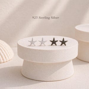 Op de afbeelding: Twee paar zeestervormige oorknopjes gemaakt van 925 sterling zilver. Eén paar is gepolijst zilver en het andere paar is donker van kleur. De tekst 925 Sterling Silver is zichtbaar boven de oorbellen, die op een ronde standaard staan.