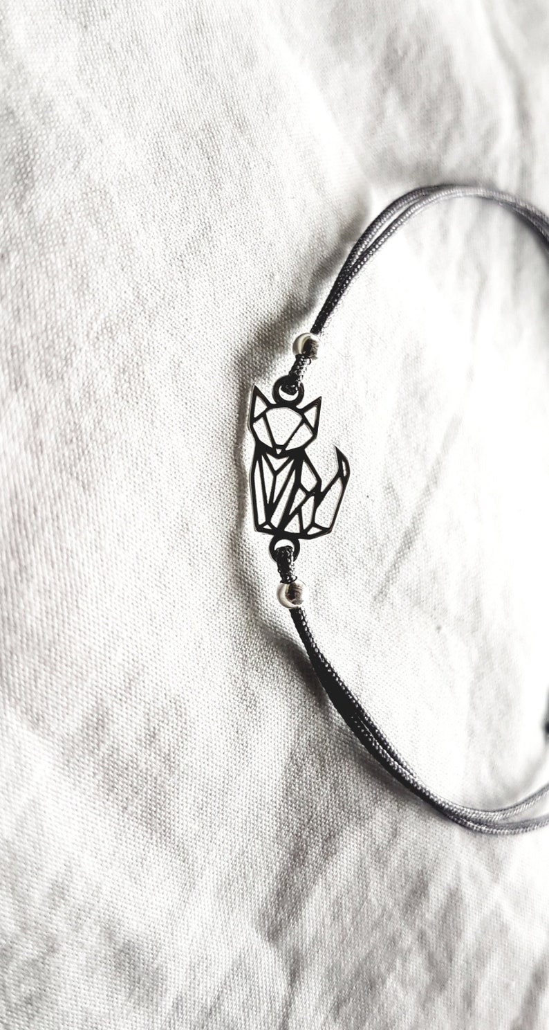 May include: A black cord bracelet featuring a central geometric fox charm made of thin black metal lines. The charm is flanked by two small silver-toned metallic beads on the cord, which is composed of two thin strands of black string.