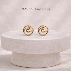 Könnte beinhalten: Ein Paar goldfarbene Ohrstecker mit einem kreisförmigen Rahmen und einem wellenförmigen Ausschnitt. Der Text 925 Sterling Silver ist über den Ohrringen zu sehen. Die Ohrringe haben eine polierte Oberfläche und ein offenes Design.