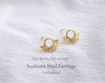 Peut inclure: Une paire de boucles d'oreilles à clous en argent sterling 925 plaqué or. Chaque boucle présente un motif circulaire central avec des rayons rayonnants s'étendant de la moitié inférieure, créant une apparence de soleil sur un fond en tissu blanc.