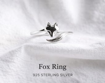 May include: A silver fox ring with a detailed fox head and tail design. The ring is made of 925 sterling silver and is displayed on a white surface. The text "Fox Ring" and "925 STERLING SILVER" are below the ring.