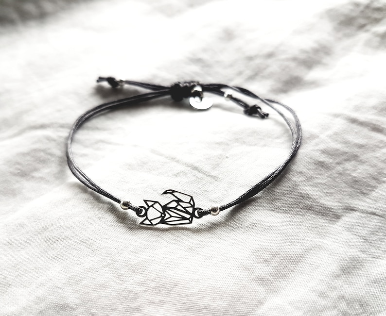 May include: A black cord bracelet featuring a central geometric fox charm made of thin black metal lines. The bracelet includes two small silver-toned metal beads flanking the fox charm and a small circular metal tag near the adjustable knot closure.