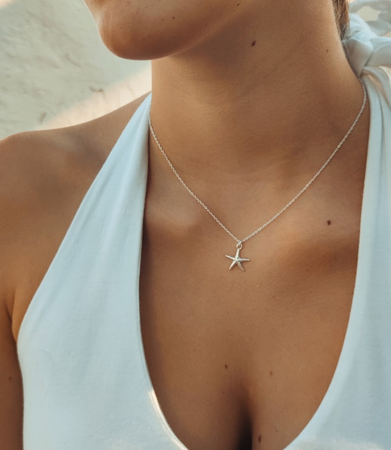 Könnte beinhalten: Eine silberne Halskette mit einem Seestern-Anhänger. Die Halskette hat eine zarte Kette und einen kleinen, fünfzackigen Seestern-Anhänger. Der Anhänger ist hellsilbern und hängt an einer dünnen Kette.