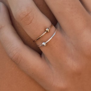 Könnte beinhalten: Zwei dünne Stapelringe, einer in Goldoptik und einer in Silberoptik, jeweils mit einer einzelnen kleinen, glatten, runden Perle in der Mitte des Rings. Die Ringe werden zusammen an einem Finger getragen und zeigen ihre polierte Metalloberfläche.