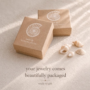 May include: Two small, square, light brown jewelry boxes with white seashell designs and the text "made by OCEANLIGHTS." Several seashells are scattered near the boxes. The text "your jewelry comes beautifully packaged ready to gift" is at the bottom.