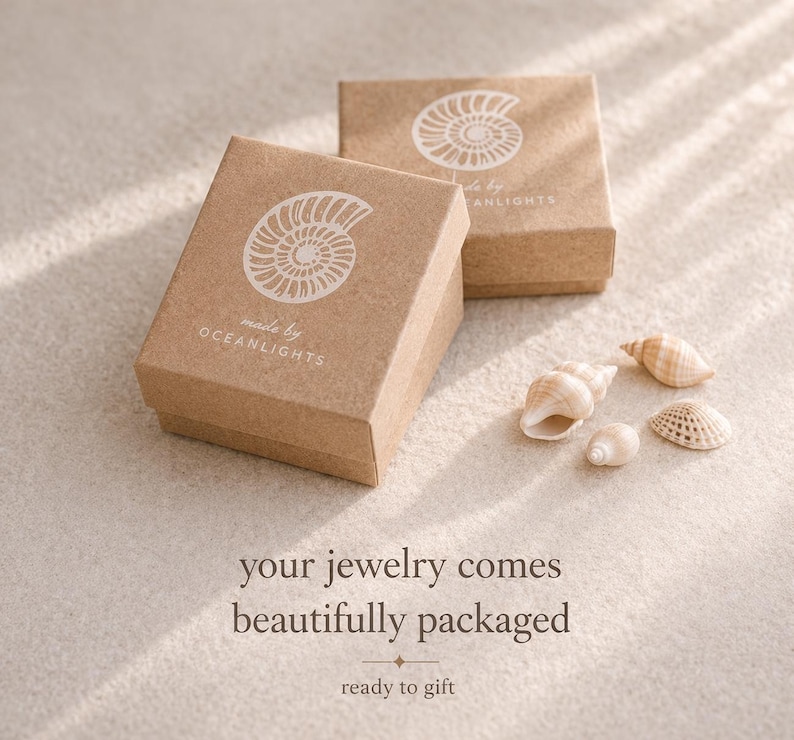 May include: Two small, square, light brown jewelry boxes with white seashell designs and the text "made by OCEANLIGHTS." Several seashells are scattered near the boxes. The text "your jewelry comes beautifully packaged ready to gift" is at the bottom.