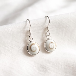 Könnte beinhalten: Ein Paar Ohrhänger mit runden, weiß-braunen spiralförmigen Shiva-Muschel-Cabochons in polierten silberfarbenen Metallfassungen. Die Ohrringe hängen an einfachen silberfarbenen Metallhaken und zeigen ein natürliches, spiralförmiges Muster.