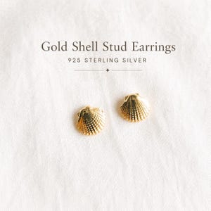 Könnte beinhalten: Goldene Muschel-Ohrstecker aus 925er Sterlingsilber. Die Ohrringe sind goldfarben und in Form kleiner Muscheln gestaltet. Sie werden auf einer weißen, strukturierten Oberfläche präsentiert. Der Text "Gold Shell Stud Earrings" und "925 STERLING SILVER" ist sichtbar.