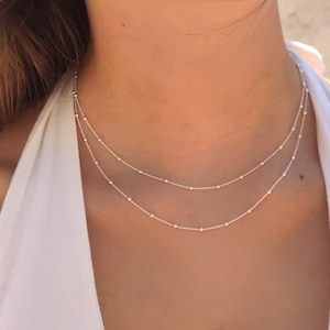 Può includere: Una collana in argento a doppio strato composta da due catenine delicate decorate con piccole perline metalliche distanziate uniformemente. Le catene sono sottili e riflettenti, poggiate sul collo in una disposizione a strati con finitura lucida.