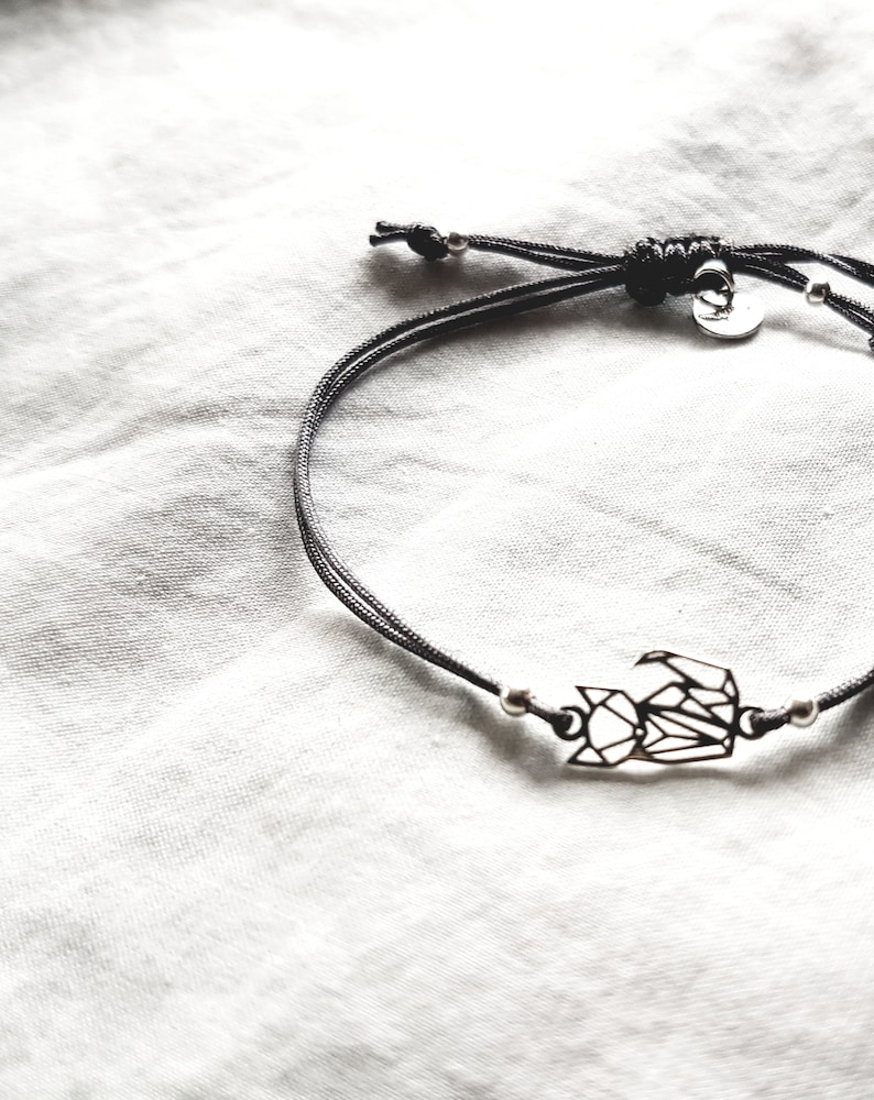 May include: A black cord bracelet featuring a silver-toned geometric fox charm. The bracelet includes small silver beads on either side of the charm and a sliding knot closure with a small circular metal tag attached near the adjustable cord ends.