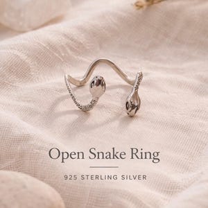 Pode incluir: Um anel de cobra aberto em prata. O anel apresenta um design ondulado com detalhes de cabeça de cobra em cada extremidade. O anel é exibido em um tecido bege. O texto "Open Snake Ring" e "925 STERLING SILVER" são visíveis.