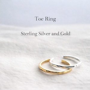 Può includere: Due anelli per le dita dei piedi, uno dorato e uno argentato, sono esposti su una superficie bianca e strutturata. L'anello dorato ha una texture martellata, mentre l'anello argentato ha una finitura liscia e lucida. Testo: "Anello per dita" e "Argento Sterling e Oro."