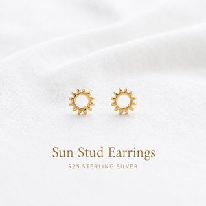 Puede incluir: Un par de pendientes de botón en forma de sol, de color dorado, fabricados en plata de ley 925. Cada pendiente presenta un centro circular rodeado de puntas radiantes. El texto Sun Stud Earrings 925 STERLING SILVER es visible.