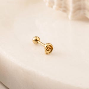 Könnte beinhalten: Ein goldfarbenes Barbell-Piercing mit einer kleinen, polierten Kugel an einem Ende und einem flachen, runden Anhänger am anderen Ende, der mit einem detaillierten, mittigen Spiralmuster versehen ist, das an eine Muschel erinnert.