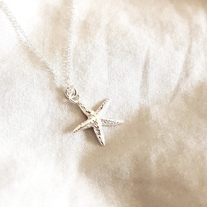May include: A silver starfish pendant necklace. The pendant is a detailed starfish design, suspended from a delicate silver chain. The necklace is displayed on a white, textured background.