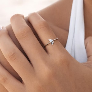 May include: A thin silver-toned ring featuring a small, delicate whale tail charm. The band has a subtle twisted or rope-like texture, and the metallic finish reflects light, highlighting the smooth, curved shape of the tail ornament.
