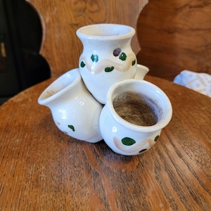 May include: A cluster of three small, off-white ceramic pots joined together. Each pot features a hand-painted design consisting of a wavy tan line accented with small, dark green dots. The interior of each pot is unglazed and shows a natural clay texture.