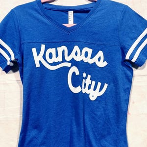 May include: A royal blue v-neck t-shirt with white stripes on the sleeves and the words "Kansas City" in white script across the chest. The shirt is hanging on a pink hanger.