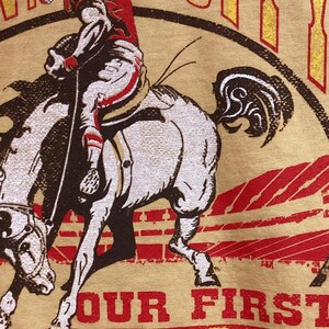 May include: Tan t-shirt featuring a rodeo scene with a rider on a bucking bronco. The rider is wearing a red helmet and outfit. The text "OUR FIRST RODEO" is printed in red and yellow.