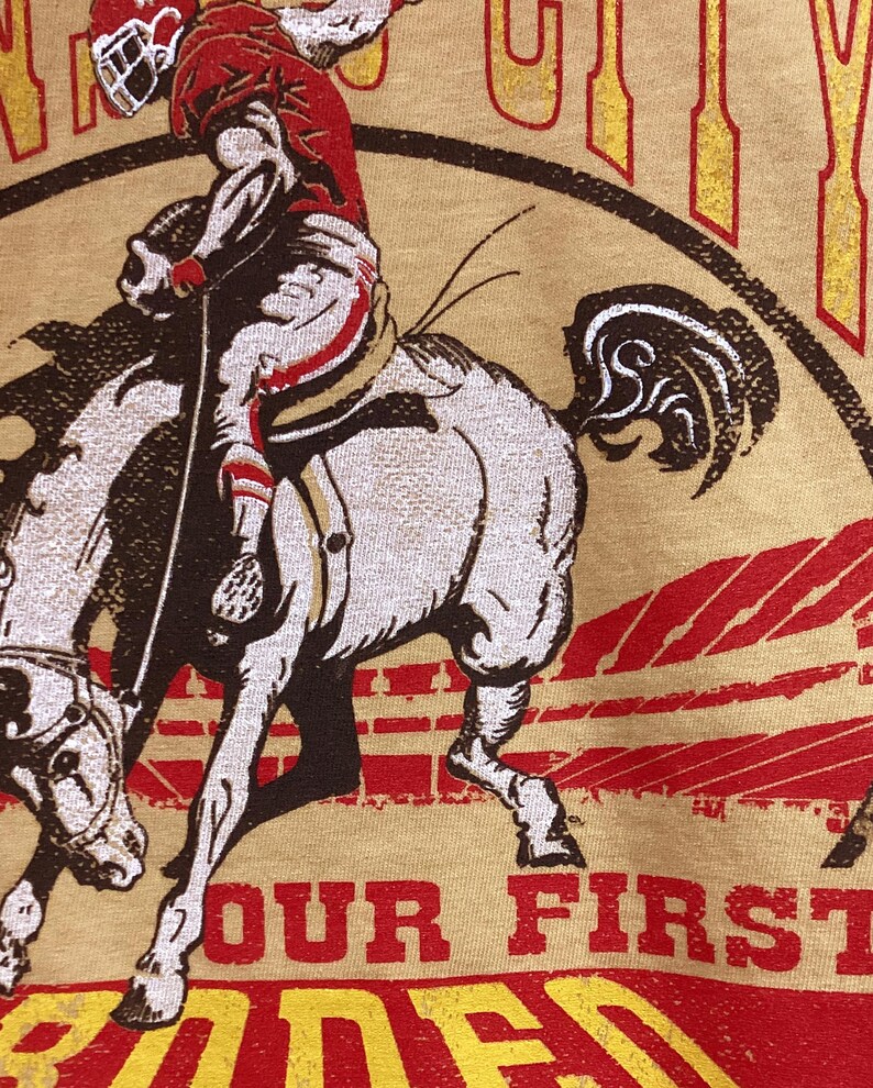 May include: Tan t-shirt featuring a rodeo scene with a rider on a bucking bronco. The rider is wearing a red helmet and outfit. The text "OUR FIRST RODEO" is printed in red and yellow.