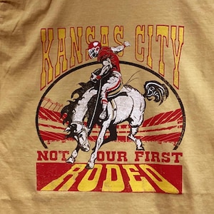 May include: Mustard yellow t-shirt with a graphic of a rodeo rider on a white horse. The text "Kansas City" is arched above the image, with "Not Our First Rodeo" below. The design is in red and yellow.