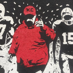 May include: Black t-shirt featuring a red graphic of a football coach wearing a KC hat, with a mustache, and pointing. Two football players are on either side of the coach. White confetti is falling from the top.