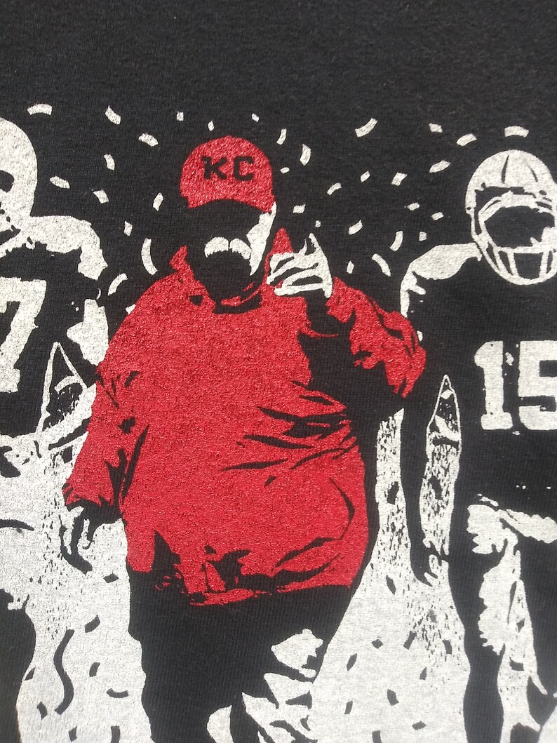 May include: Black t-shirt featuring a red graphic of a football coach wearing a KC hat, with a mustache, and pointing. Two football players are on either side of the coach. White confetti is falling from the top.