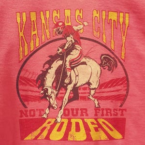 May include: Pink t-shirt with a distressed graphic featuring a football player riding a horse. The text "KANSAS CITY" arches above the image, with "NOT OUR FIRST RODEO" below in a yellow and red color scheme.