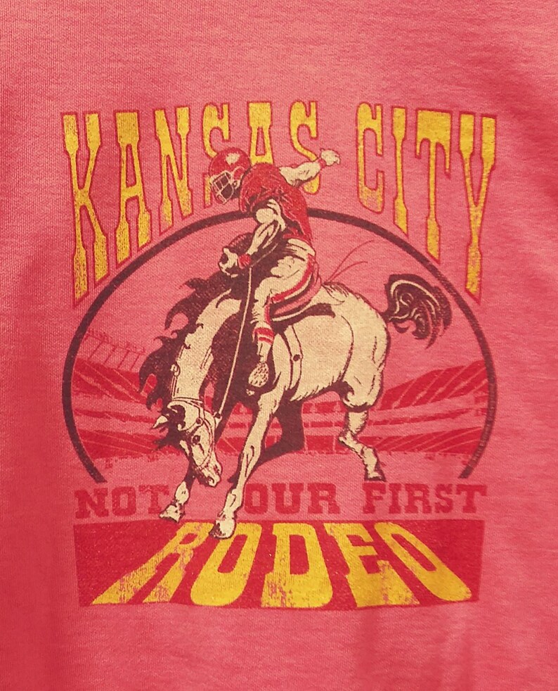 May include: Pink t-shirt with a distressed graphic featuring a football player riding a horse. The text "KANSAS CITY" arches above the image, with "NOT OUR FIRST RODEO" below in a yellow and red color scheme.