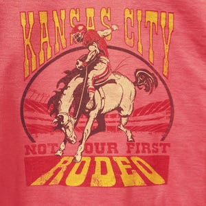 May include: Pink t-shirt featuring a graphic of a football player riding a bucking bronco. The text "Kansas City" arches above the image, with "Not Our First Rodeo" below. The design is in shades of red and yellow.