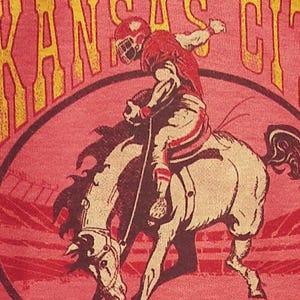 May include: A red t-shirt featuring a football player riding a bucking bronco. The player is wearing a red helmet and uniform. The words "KANSAS CITY" are arched above the image in yellow lettering. The shirt has a pink background.