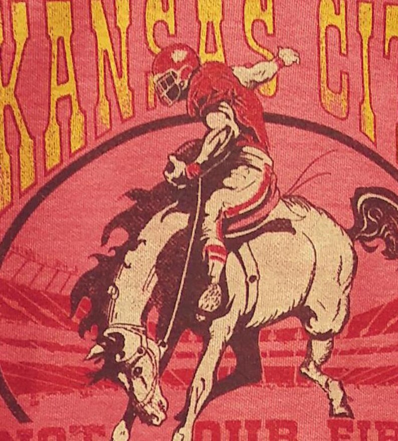 May include: A red t-shirt featuring a football player riding a bucking bronco. The player is wearing a red helmet and uniform. The words "KANSAS CITY" are arched above the image in yellow lettering. The shirt has a pink background.