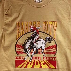 May include: Mustard-colored t-shirt featuring a graphic of a football player riding a bucking bronco with the words "Kansas City" and "Not Our First Rodeo" in red and yellow lettering. The shirt has a crew neck.