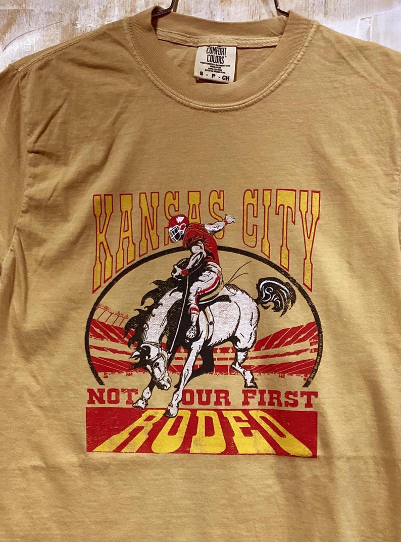 May include: Mustard-colored t-shirt featuring a graphic of a football player riding a bucking bronco with the words "Kansas City" and "Not Our First Rodeo" in red and yellow lettering. The shirt has a crew neck.